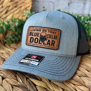 May include: A gray trucker hat with a black mesh back and a curved brim. The front features a brown leather patch with the words "LIVING ON THAT BLUE COLLAR DOLLAR" and a skull graphic. The hat has an adjustable strap.
