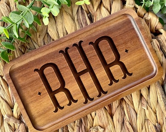 Op de afbeelding: Een rechthoekig houten dienblad met de gegraveerde letters "RHR" in een schreeflettertype. De lade heeft een warme bruine kleur en heeft een opstaande rand. Kleine stervormige details staan op de lade. De lade staat op een geweven oppervlak met groen gebladerte.
