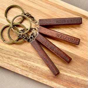 May include: A collection of wooden keychains with bronze-colored key rings. Each rectangular keychain is engraved with a different message: "RIDER & RIDGE", "WELCOME", "I LOVE YOU MORE", and "FRONT DOOR". The keychains are arranged on a wooden tray.