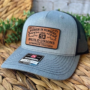 Peut inclure: Casquette de camionneur grise avec un dos en filet noir et une visière incurvée. Le chapeau présente un patch en cuir marron avec le texte "POWER IN NUMBERS LOCAL 613 BUILD UNIONS STRONGER TOGETHER". Une étiquette noire et blanche est sur la visière.