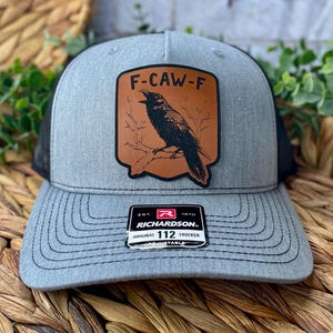 May include: Gray trucker hat with a black mesh back and a curved brim. The front features a brown leather patch with a black raven illustration and the text "F-CAW-F". The hat includes a Richardson patch.