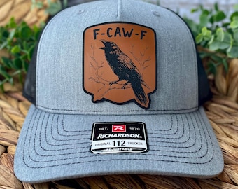 May include: Gray trucker hat with a black mesh back and a curved brim. The front features a brown leather patch with a black raven illustration and the text "F-CAW-F". The hat includes a Richardson patch.