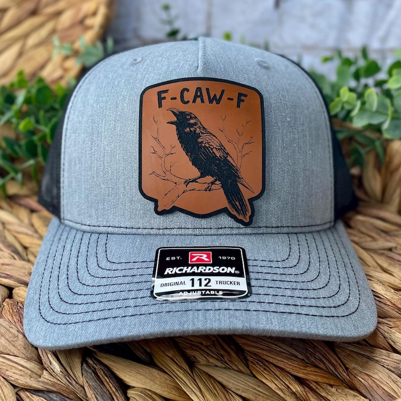 May include: Gray trucker hat with a black mesh back and a curved brim. The front features a brown leather patch with a black raven illustration and the text "F-CAW-F". The hat includes a Richardson patch.