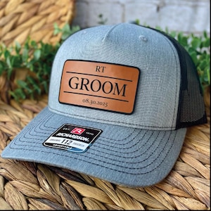 Può includere: Cappellino trucker grigio con retro in rete nera e toppa in pelle marrone. La toppa recita "RT GROOM 08.30.2025". Il cappellino ha una visiera curva e un cinturino regolabile. Il cappellino trucker Richardson 112 è regolabile.