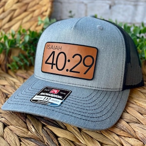 May include: Gray trucker hat with a black mesh back and a curved brim. The hat features a brown leather patch with the text "ISAIAH 40:29" in black. The hat also has a black and white label that says "RICHARDSON".
