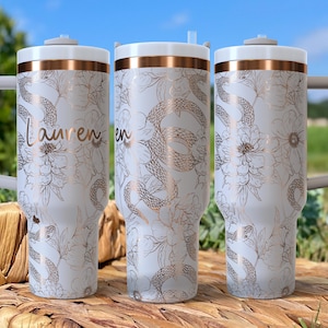 Op de afbeelding: Drie witte geïsoleerde bekers met een bloemen- en slangenontwerp in goud. De bekers hebben een wit deksel en een gouden band rond de bovenkant. De naam "Lauren" staat op een van de bekers.