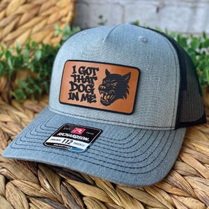May include: Gray trucker hat with a black mesh back and a curved brim. The hat features a brown leather patch with the text "I GOT THAT DOG IN ME" and a graphic of a snarling dog. The hat is adjustable.