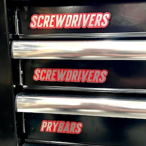 May include: A black tool chest with three drawers. The top two drawers are labelled "SCREWDRIVERS" in red and silver lettering. The bottom drawer is labelled "PRYBARS" in red and silver lettering. The drawer pulls are silver.