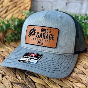 May include: A grey trucker hat with a black mesh back. The hat features a brown leather patch with the text "GREG'S GARAGE ESTABLISHED 2016" and a wrench and gear graphic. The hat is adjustable.