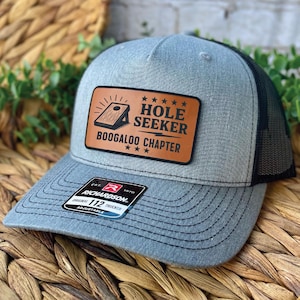May include: Gray trucker hat with a black mesh back and a curved brim. The hat features a brown leather patch with the words "HOLE SEEKER BOOGALOO CHAPTER" and a cornhole graphic. The Richardson brand tag is visible.