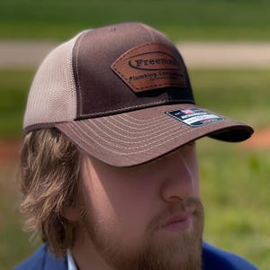 A man wearing a custom trucker hat featuring his business logo engraved on a leatherette patch, showcasing the fit and look when worn.