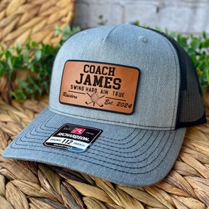 Pode incluir: Boné de caminhoneiro cinza e preto com um patch de couro que diz "COACH JAMES SWING HARD. AIM TRUE Raiders Est. 2024". O boné é ajustável e tem uma etiqueta Richardson.