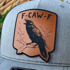 May include: A gray baseball cap with a brown leather patch featuring a black raven perched on a branch. The patch has the text "F-CAW-F" in white letters. The cap has a curved brim and a structured crown.