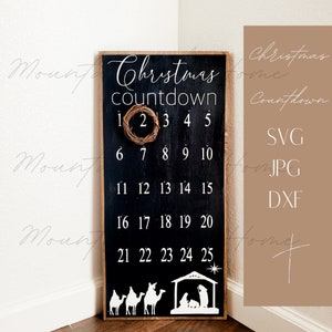 May include: Black wooden sign with white text that says "Christmas countdown" and numbers 1-25. A small wreath is hanging on the number 2. The sign also has a white silhouette of a nativity scene with three camels.