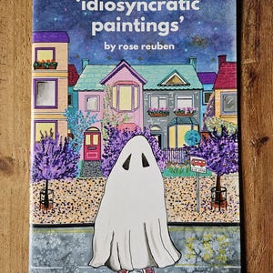 May include: A book cover with the title "'idiosyncratic paintings' by rose reuben". The cover features a colourful illustration of a street scene with houses, a ghost, and a bus stop.