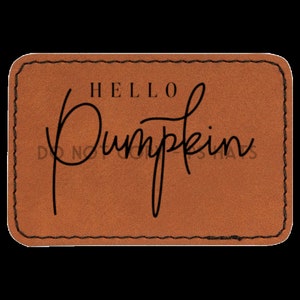 Puede incluir: Un parche de cuero marrón con un borde cosido. El texto "HELLO PUMPKIN" está escrito en letra cursiva negra.