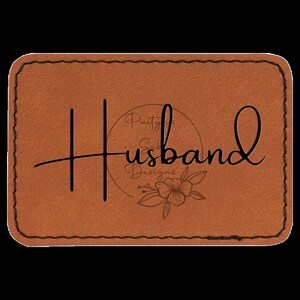 Op de afbeelding: Bruin leren patch met een gestikte rand. De tekst "Husband" is geschreven in een cursief schrift met een cirkel die de tekst "Paity's Sweet Designs" bevat en een bloemmotief eronder.