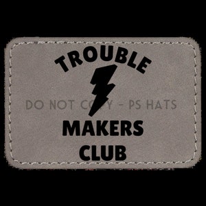 May include: A gray patch with black stitching around the edges. The patch has black text that reads "Trouble Makers Club" with a lightning bolt graphic.
