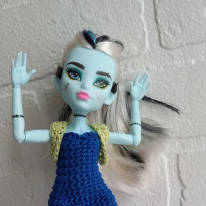 Könnte beinhalten: Eine blaue und grüne Puppe mit weißen Haaren und einem blauen Kleid. Die Puppe hat eine genähte Narbe am Hals und ein silbernes Prothesenbein mit dem Wort "FRANK" darauf.