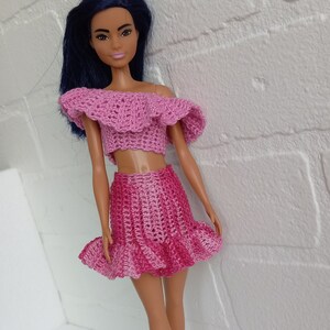 Könnte beinhalten: Eine Puppe, die ein rosafarbenes, gehäkeltes Zweiteiler-Outfit mit einem Rüschenoberteil und einem Rock trägt. Die Puppe hat langes schwarzes Haar und trägt rosa Schuhe.