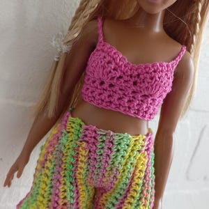 May include: A doll wearing a pink crocheted crop top and multi-colored crocheted shorts.