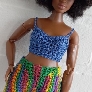 May include: A doll wearing a blue crocheted crop top and rainbow crocheted shorts. The doll has brown skin and a large afro.