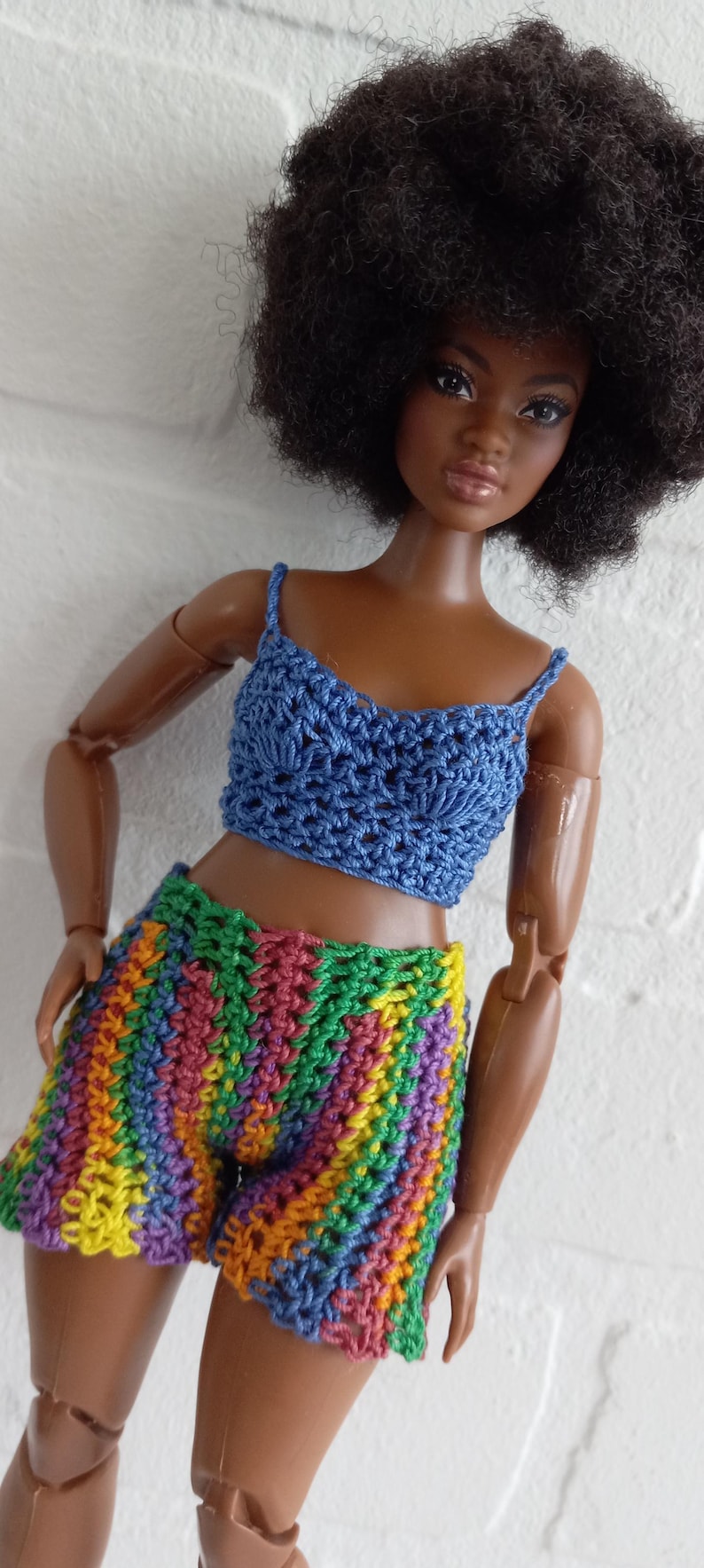 May include: A doll wearing a blue crocheted crop top and rainbow crocheted shorts. The doll has brown skin and a large afro.