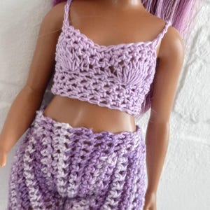 May include: A purple crochet top and shorts set for a doll. The top has a scalloped neckline and the shorts have a striped pattern.