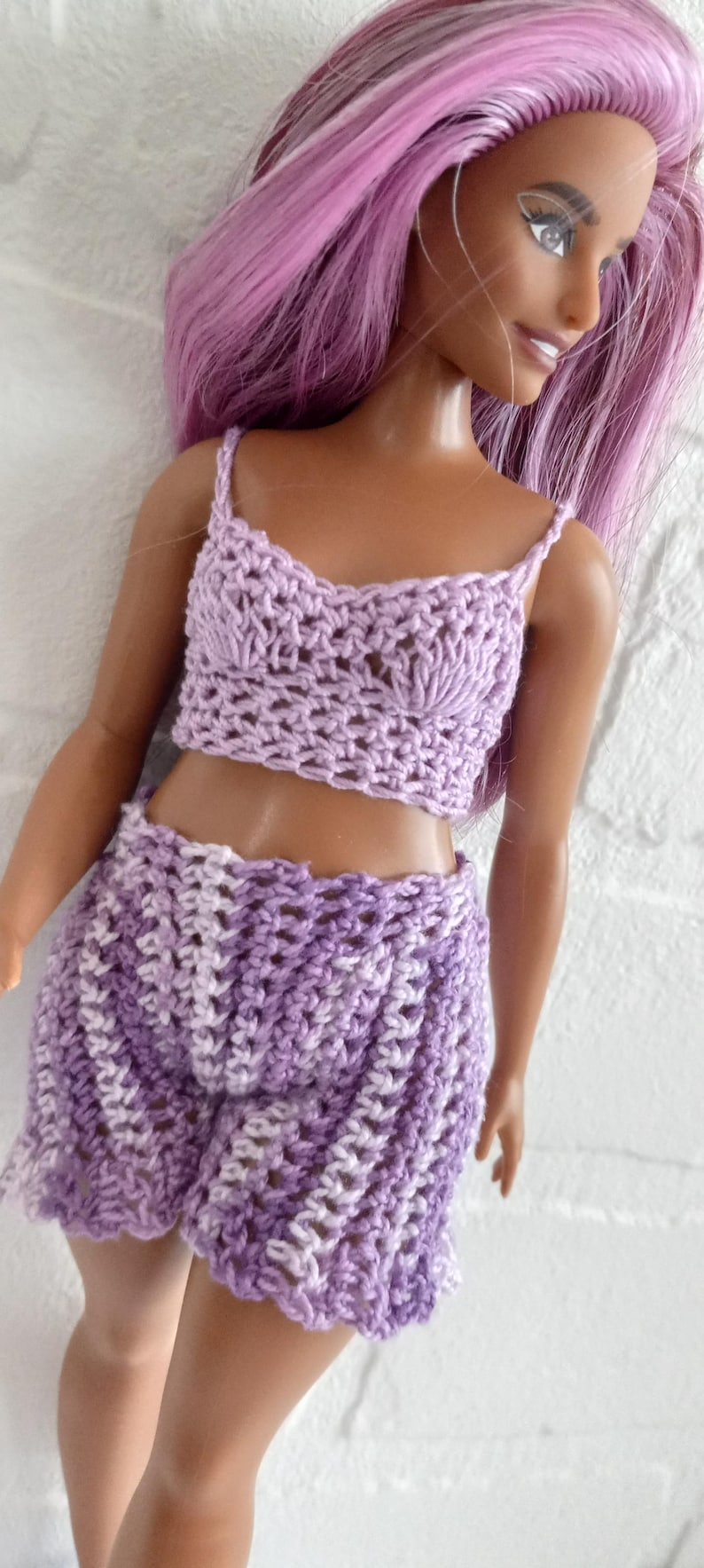 May include: A purple crochet top and shorts set for a doll. The top has a scalloped neckline and the shorts have a striped pattern.