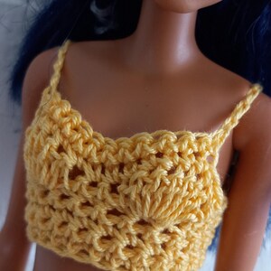 May include: A doll wearing a yellow crocheted crop top and a multi-colored crocheted skirt.