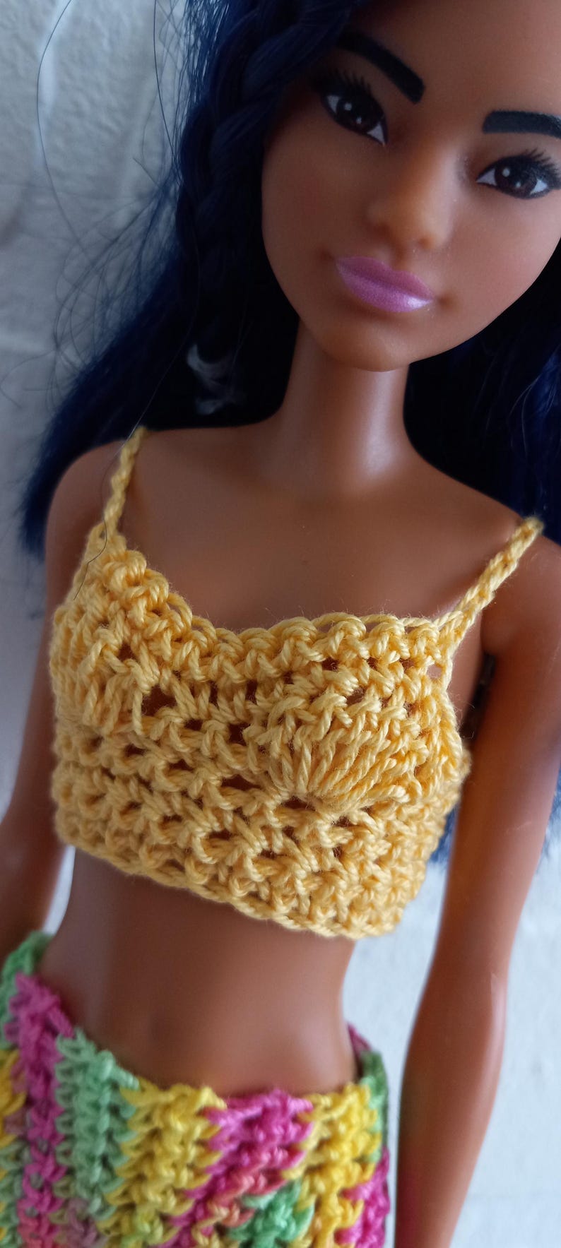 May include: A doll wearing a yellow crocheted crop top and a multi-colored crocheted skirt.