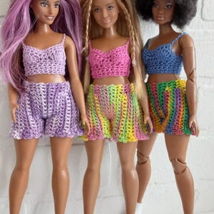 May include: Three dolls wearing crocheted outfits. The doll on the left is wearing a purple top and shorts. The doll in the middle is wearing a pink top and green and yellow striped shorts. The doll on the right is wearing a blue top and rainbow striped shorts.