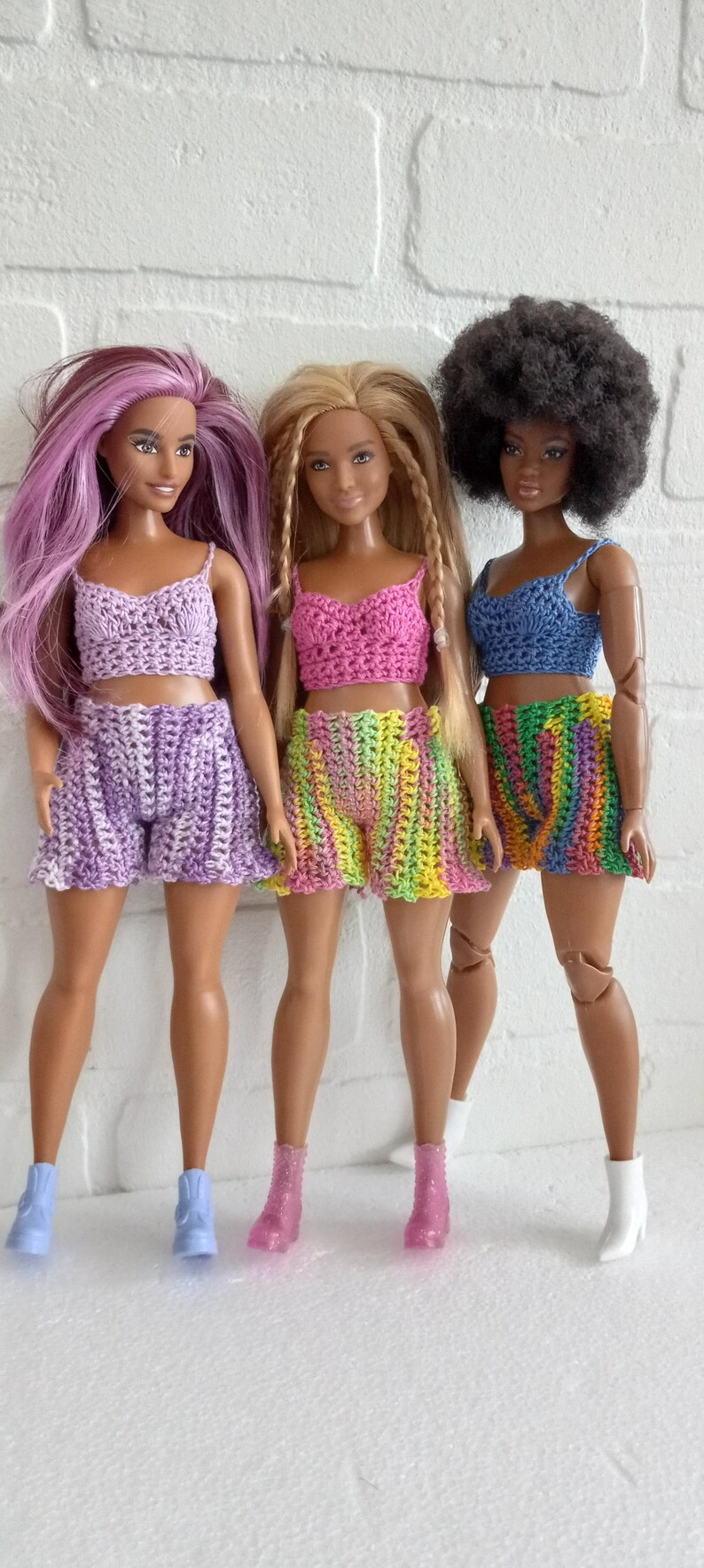 May include: Three dolls wearing crocheted outfits. The doll on the left is wearing a purple top and shorts. The doll in the middle is wearing a pink top and green and yellow striped shorts. The doll on the right is wearing a blue top and rainbow striped shorts.