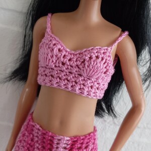 May include: A pink crocheted two-piece outfit for a doll. The top is a halter style with a scalloped neckline and the skirt is a mini skirt with a scalloped hem.