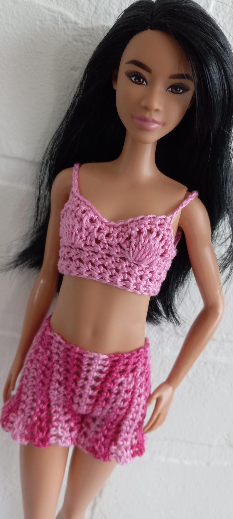 May include: A pink crocheted two-piece outfit for a doll. The top is a halter style with a scalloped neckline and the skirt is a mini skirt with a scalloped hem.