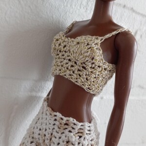 May include: A doll wearing a white crocheted top and shorts with gold accents. The doll has dark brown skin and black curly hair.