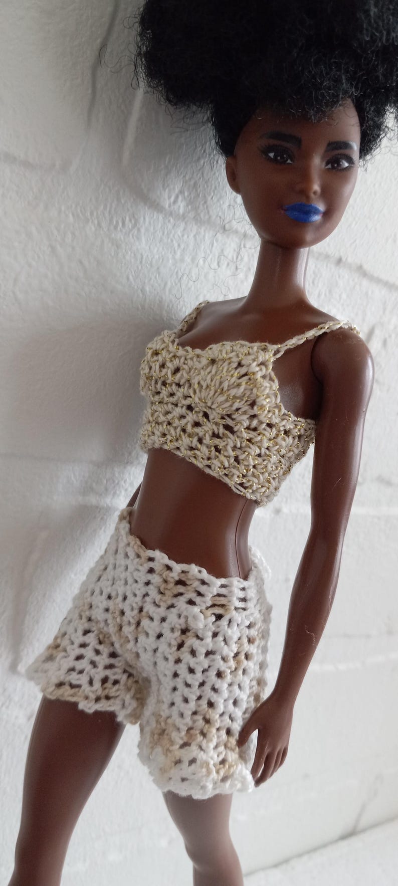 May include: A doll wearing a white crocheted top and shorts with gold accents. The doll has dark brown skin and black curly hair.
