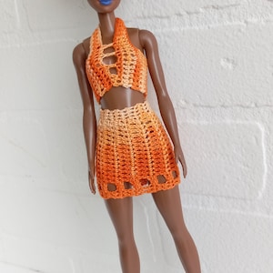 May include: A doll with black afro hair wearing a crocheted orange and white halter top and skirt. The doll is wearing lavender shoes.