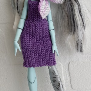 Könnte beinhalten: Eine blaue und weiße Puppe mit schwarz-weißem Haar, die ein lila Kleid und rosa Turnschuhe trägt. Die Puppe hat ein silbernes Prothesenbein mit dem Wort "Frankie" darauf.