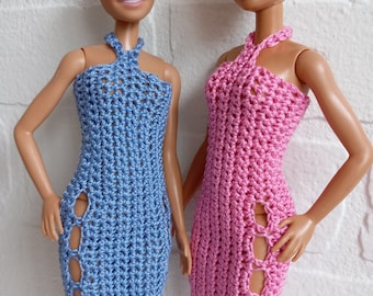 May include: Two bald-headed dolls wearing crocheted dresses. The dress on the left is blue and the dress on the right is pink. Both dresses have a halter neckline and a side slit.