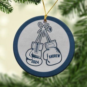 May include: A blue and white ceramic Christmas ornament with a pair of boxing gloves tied together with a ribbon. The gloves are white with blue trim and the text "ANDREW" and "2024" are printed on them.