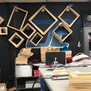 May include: A workshop with wooden picture frames hanging on a black wall. The frames are various sizes and shapes. There are stacks of wooden frames on a table with a staple gun and other tools.
