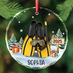 May include: A clear, round Christmas ornament featuring a pair of yellow and black swim fins, a snorkel, and a diving mask. The ornament has the name "SOPHIA" and the year "2025" on it. The background includes snow, trees, and gifts.