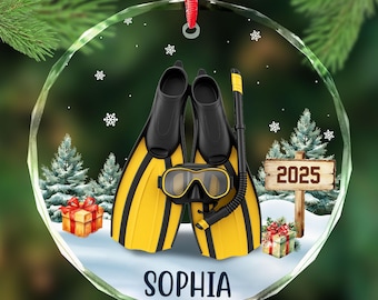 May include: A clear, round Christmas ornament featuring a pair of yellow and black swim fins, a snorkel, and a diving mask. The ornament has the name "SOPHIA" and the year "2025" on it. The background includes snow, trees, and gifts.