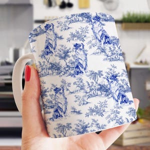 May include: White ceramic mug featuring a blue and white toile design with a repeating pattern of dogs in a landscape setting. The mug has a curved handle and a glossy finish. The design is reminiscent of classic porcelain.
