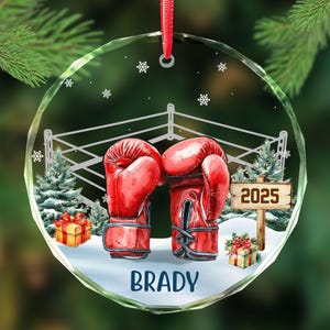 May include: A clear, round Christmas ornament with a boxing theme. Two red boxing gloves face each other in front of a boxing ring. The name "BRADY" is at the bottom, with "2025" on a sign. Snowflakes and Christmas trees complete the design.