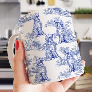 Może przedstawiać: Biały ceramiczny kubek z niebieskim wzorem toile de Jouy przedstawiającym Golden Retrievery i liście. Wzór powtarza się wokół kubka, tworząc klasyczną estetykę. Kubek ma biały uchwyt.