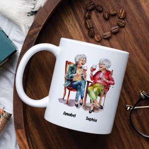 Puede incluir: Taza de cer&aacute;mica blanca con una ilustraci&oacute;n de acuarela de dos mujeres mayores sentadas en sillas y sosteniendo copas de vino. El texto "Amabel" y "Sophia" est&aacute; impreso debajo de la imagen.