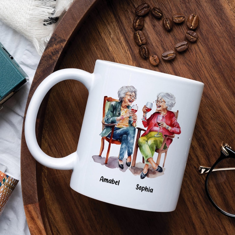 Puede incluir: Taza de cer&aacute;mica blanca con una ilustraci&oacute;n de acuarela de dos mujeres mayores sentadas en sillas y sosteniendo copas de vino. El texto "Amabel" y "Sophia" est&aacute; impreso debajo de la imagen.