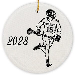 May include: A black and white illustration of a lacrosse player wearing a helmet and jersey with the number 15 and the name "BRADY" on it. The player is holding a lacrosse stick and is running with the ball. The year 2023 is written in black cursive on the left side of the image.